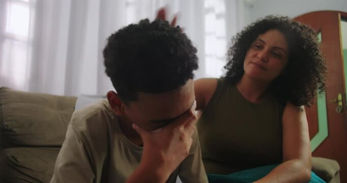Teenager boy sitting on couch, emotionally struggling and covering his face with hand, mother showing support with a comforting gesture, family empathy and care