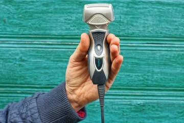 one electric safe with sharp iron blades and protective grid with battery charge indicators and power button plastic compact gray with black cord men's razor in hand near wooden green wall