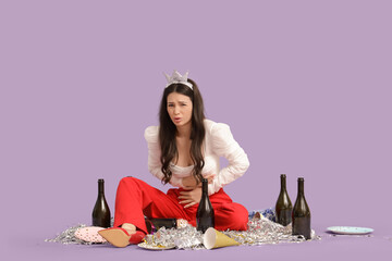 Young woman with bottles feeling unwell after party on lilac background