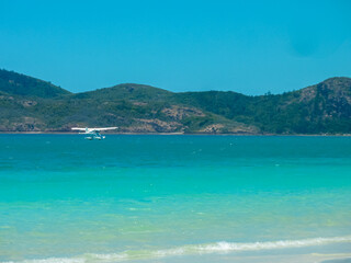 Seaplane glides low over the crystal-clear turquoise waters of the Whitsunday Islands, Queensland, Australia. White aircraft contrasts against mountainous green islands. Pristine azure waters