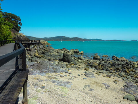 Wooden boardwalk winding along rocky coastline at Airlie Beach, Queensland, Australia. Turquoise waters of Coral Sea with sailboats in distance, against backdrop of green hills under clear blue sky - Powered by Adobe