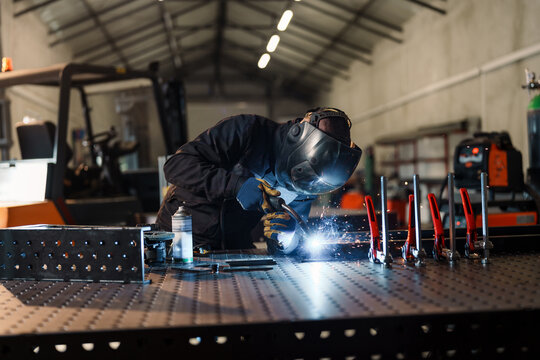 Professional welder in protective gear using specialized equipment to join metal pieces, creating sparks in a modern industrial setting with a forklift nearby