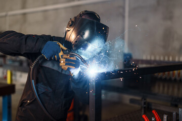 Professional welder wearing protective gear and using specialized equipment is joining metal profiles, creating a shower of sparks and smoke in a workshop environment