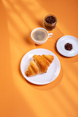 Tasty french croissant on a white plate and a cup of americano coffee in sunlight rays from above on an orange background. Sweet breakfast. Morning food.