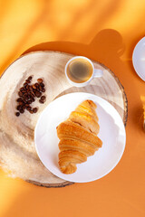 Tasty french croissant on a white plate and a cup of americano coffee in sunlight rays from above on an orange background. Sweet breakfast. Morning food.