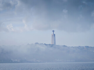 Cristo Rei Statue in the fog next to 25 April Bridge near Tagus river. Sanctuary of Christ the King...