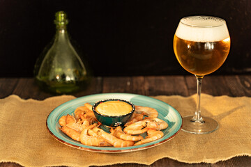 A glass of light beer and large shrimps and sauce on a turquoise plate on a wooden table. Selected focus.