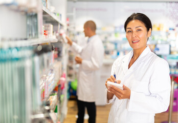 Obraz premium Positive female pharmacist makes notes on a notebook in the sales area of a pharmacy