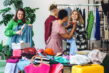 Cheerful young student friends having fun at home swap party, exchanging clothes, trying hats. Eco conscious lifestyle, ecology protection. Preventing textile pollution. Sustainable fashion concept