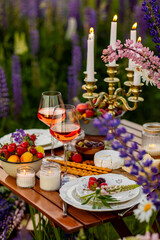 Two glasses of rose wine, flowers in a vase, silverware, fruits. Sunset, golden hour, atmospheric candlelight. Romantic table decor for a loving couple on blooming meadow with purple lupines