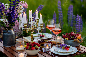 Two glasses of rose wine, flowers in a vase, silverware, fruits. Sunset, golden hour, atmospheric candlelight. Romantic table decor for a loving couple on blooming meadow with purple lupines