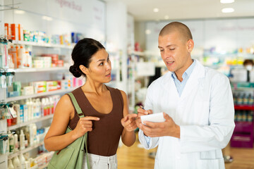 Obraz premium Male pharmacist in white coat writing recipe for Asian woman while standing in salesroom of drugstore.