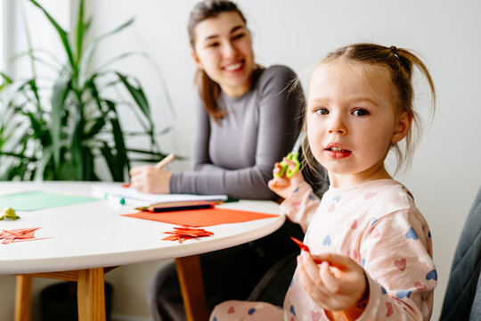 Girl, 3 years old, and mother do arts and crafts together, cutting shapes from colored paper while sitting in kitchen at home. Motherhood, parenting, and educational activities concept.