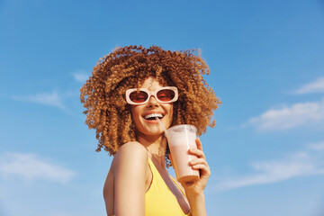 Happy woman with curly hair enjoying a smoothie outdoors against a blue sky