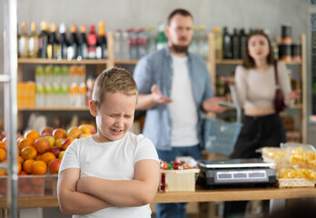 Capricious preteen boy standing with crossed arms and screaming loudly in grocery store while frustrated puzzled parents looking at son in background ..