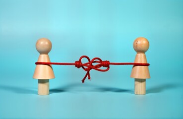 Two wooden figurines tied together by red rope with knot in middle. Figurines represent couple facing difficulties in relationships. Figurines bound together conflict, stress. Image shows complex