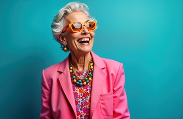 Stylish senior woman with vibrant pink blazer, colorful necklace. Wears trendy sunglasses, joyful expression. Appears happy, active. Cool grandmother embodies fashionable aging. Trendsetter in trendy