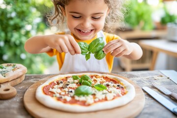 Happy little girl adding fresh basil leaves to a homemade pizza, enjoying cooking in the kitchen