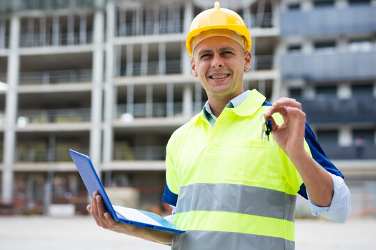 Portrait of a positive man engineer on a construction site, demonstrating in his hand the keys to a flat in a new building