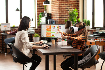 Woman uses internet for research on desktop pc as daughter wears vr glasses to experience artificial intelligence. Mother reviews data charts on computer and keeps her child entertained with 3d