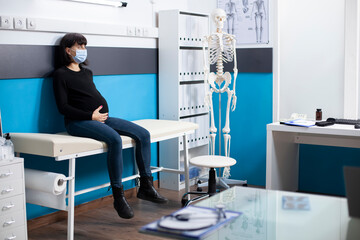 Expectant mother in face mask gently holds her pregnancy belly, awaiting checkup visit in clinic room filled with medical tools. Pregnant woman seated on hospital bed ready for health examination.