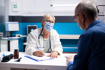 Elderly physician discusses healthcare treatment options with senior white male patient seated in clinic office. Retired doctor attentively listening to concerns of old man and writing on clipboard.