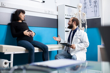 Male obstetrician uses digital device to discuss pregnancy care details with young expectant mother during checkup. Friendly physician with tablet, giving prenatal treatment plans to pregnant patient.