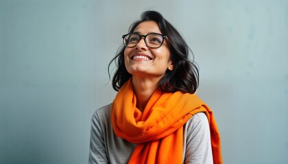 Happy Indian woman wearing eyeglasses smiles looking upwards against gray background. Stylish woman wears orange scarf. Portrait shows positive emotion, style. Image shows fashion, health. Great for