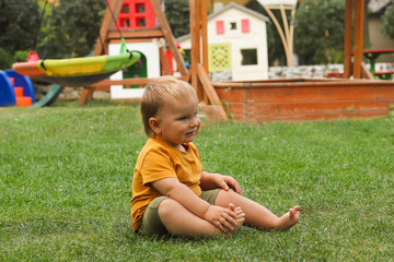 A cheerful toddler sitting on green grass in an outdoor playground. The child is wearing an orange T-shirt and olive-green shorts, smiling happily while looking to the side