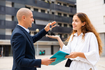 Man real estate manager giving keys and paper-folder with documentation to woman who have purchased apartment in new multistory house.