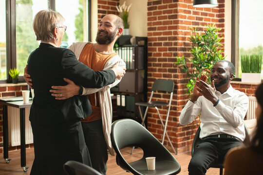 Retired female counselor warmly hugs male patient at aa meeting, applauding his successful recovery from addiction. Elderly psychiatrist and black man encouraging bearded guy at group therapy session.