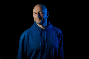Studio portrait of a bald man with grey beard on dark background. Male in blue hoodie illuminated by warm and cool blue light. Simple comfortable clothes. Smile on face. Calm and confident person.