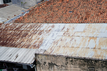A high-angle view of old rooftops, showing a mix of rusty corrugated metal and aged clay tiles. The scene depicts urban decay. Rusty and Clay Rooftops in Urban Setting
