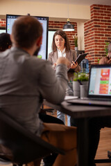 White woman with clipboard in hand, presenting business plan with her coworkers in startup office. Caucasian female manager standing in center of room and speaking about marketing strategy with team.