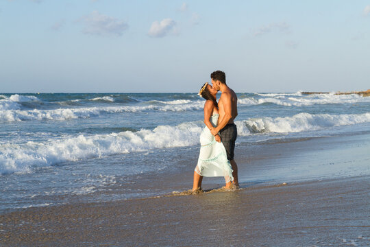 Loving young couple at the beach in a late summer hazy day at dusk, wearing a turquoise dress and shorts, enjoying going barefoot in the ocean water, getting wet, teasing and kissing one another.