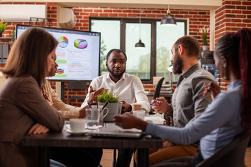 Group of diverse colleagues sitting around table and holding business meeting in brick wall room. African american manager leads conversation with coworkers, discussing project development of company.