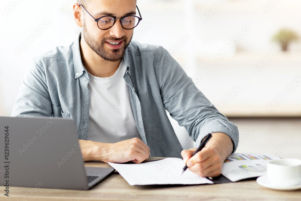 Poster young caucasian businessman taking notes while working on laptop computer at home office. freelancer