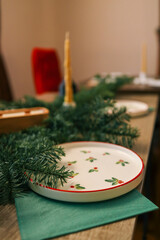 A festive table setting with a decorative ceramic plate featuring a holly pattern, placed on a green napkin. Spruce branches add a natural touch, while a candle in the background