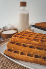 Close-up of waffle-making ingredients with a waffle maker in the background. Wooden bowls with flour and sugar, eggs in a carton, and a golden waffle resting on the machine
