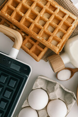 Ingredients for homemade waffles arranged on a white surface, including flour, sugar, eggs, and milk. A waffle maker with wooden handles is visible in the frame