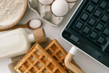 Ingredients for homemade waffles arranged on a white surface, including flour, sugar, eggs, and milk. A waffle maker with wooden handles is visible in the frame