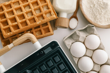 Ingredients for homemade waffles arranged on a white surface, including flour, sugar, eggs, and milk. A waffle maker with wooden handles is visible in the frame