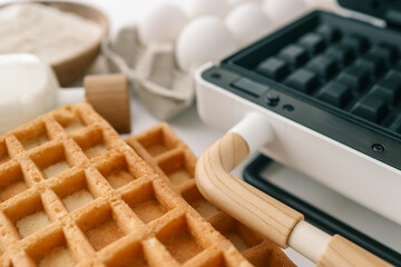 Close-up of a waffle iron with wooden handle, alongside freshly baked waffles and ingredients in a kitchen setting