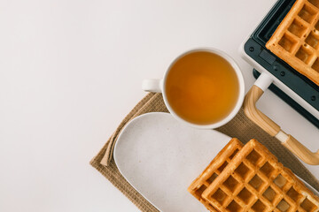 Freshly baked golden waffles served on a white plate with a glass of honey. Crisp texture and inviting aroma. A warm breakfast scene with a waffle maker in the background