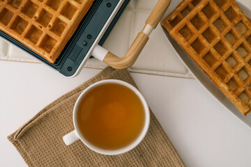 Freshly baked golden waffles served on a white plate with a glass of honey. Crisp texture and inviting aroma. A warm breakfast scene with a waffle maker in the background