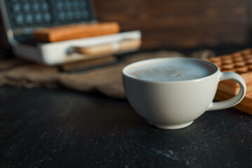 A steaming cup of coffee next to freshly made waffles on a dark textured background, with a waffle maker in the blurred background. Cozy breakfast atmosphere