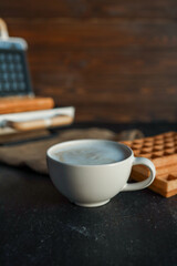 A steaming cup of coffee next to freshly made waffles on a dark textured background, with a waffle maker in the blurred background. Cozy breakfast atmosphere