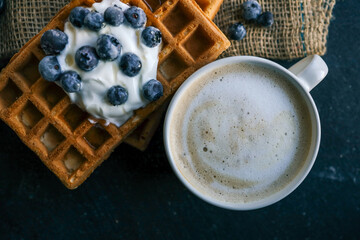 Stack of golden waffles topped with whipped cream and fresh blueberries, served on a rustic plate with a blurred cup of coffee in the background. A cozy, delicious breakfast setting