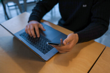 A person multitasking by holding a smartphone and typing on a laptop at a wooden desk, with large glass windows and festive decorations in the blurred background