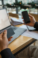 A person holding a black credit card and a smartphone in front of a laptop, making an online payment. Digital transaction, e-commerce, and online shopping concept with a modern workspace setting